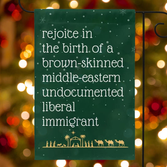 Green garden flag with snow in background and gold manger christmas scene along the bottom. Text reads in white font rejoice in the birth of a brown skinned middle eastern undocumented liberal immigrant