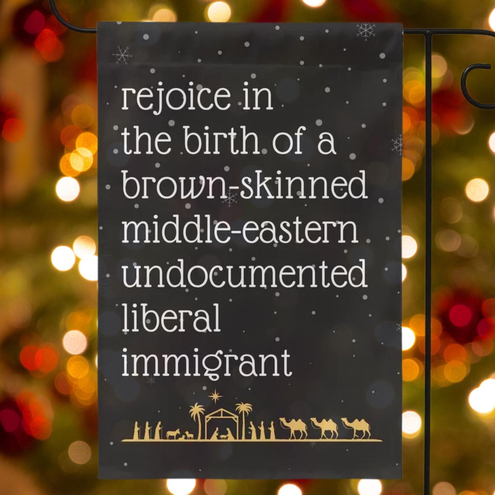 Black garden flag with snow in background and gold manger christmas scene along the bottom. Text reads in white font rejoice in the birth of a brown skinned middle eastern undocumented liberal immigrant